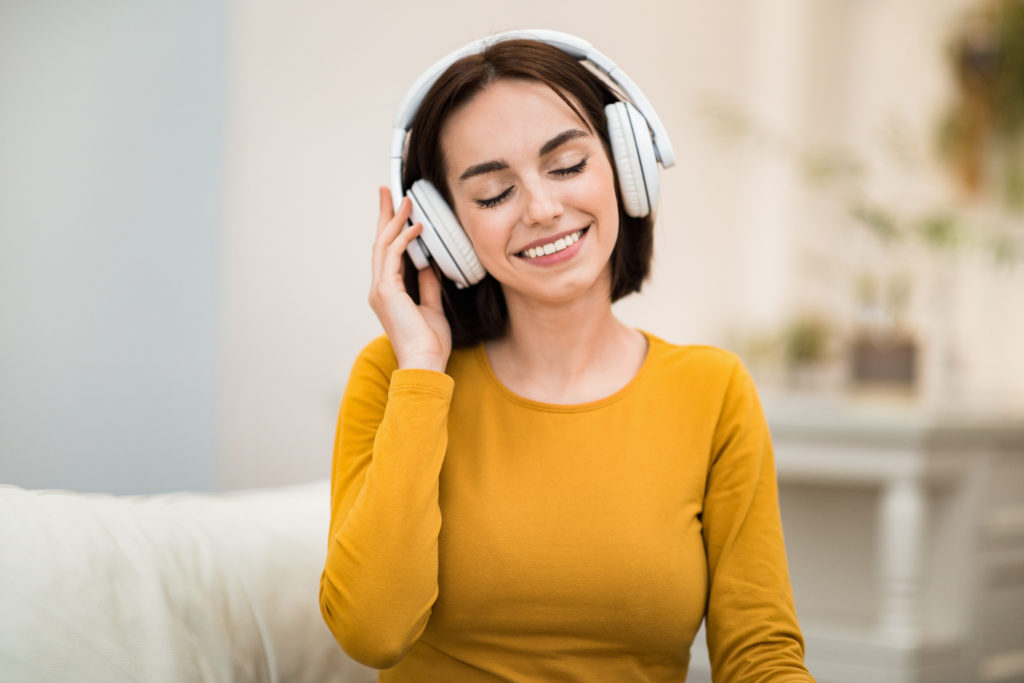 Joyful happy attractive brunette young woman sitting on couch at home, listening to music with closed eyes and smiling, lady using modern wireless stereo headphones